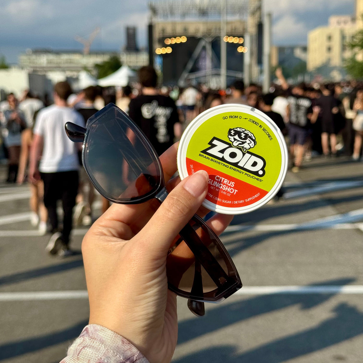 A hand holds a round tin of Zoid Citrus Slingshot in front of a blurred outdoor crowd scene with a stage in the background.