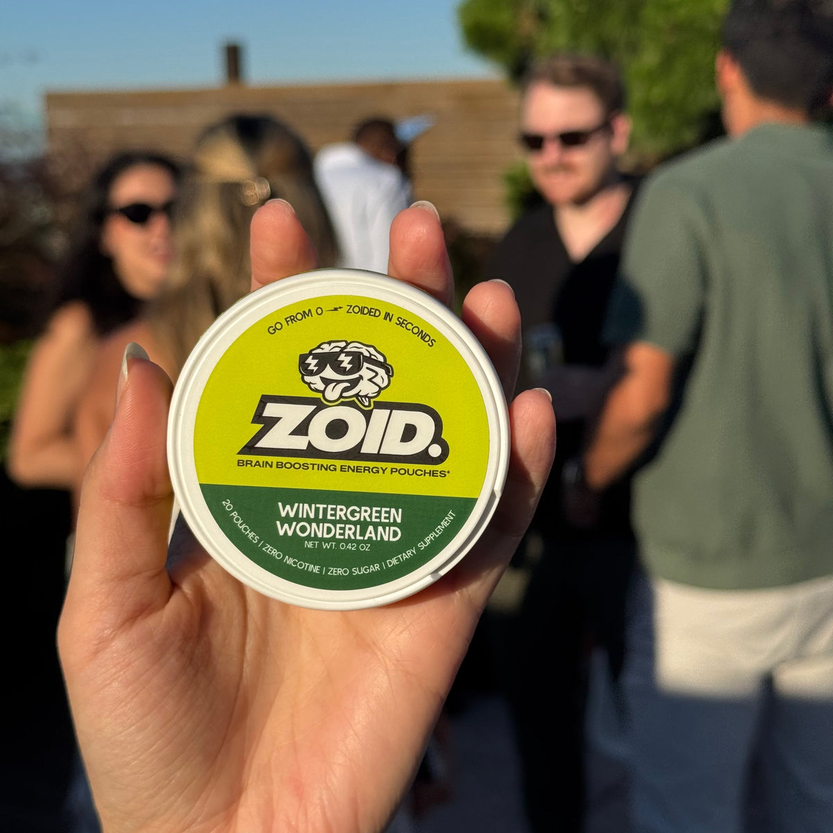 A hand holds a round tin of Zoid Brain-Boosting Energy pouches in the flavor Wintergreen Wonderland, with a blurred outdoor party background featuring people.