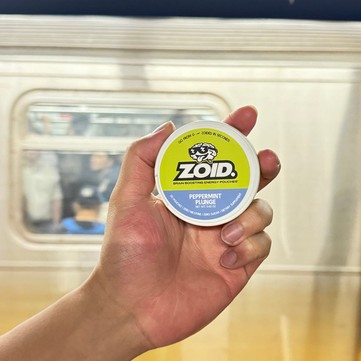 A hand holding a small round container of ZOID Peppermint Plunge in front of a blurred subway train in a station.