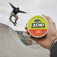 A hand holds a round tin of Zoid Brain Boosting Energy Pouches in Citrus Slingshot flavor, with a skateboarder jumping a trick in a skatepark blurred in the background.