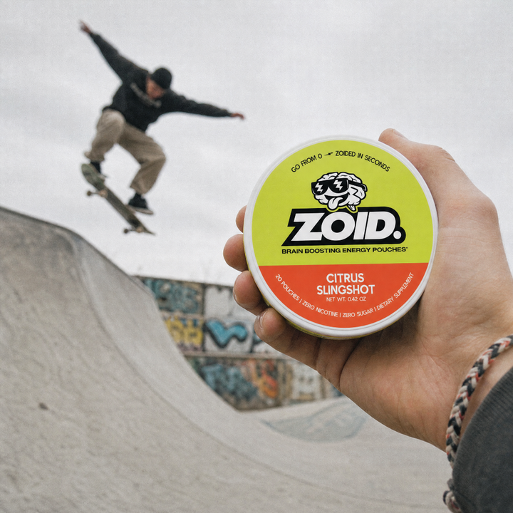 A hand holds a round tin of Zoid Brain Boosting Energy Pouches in Citrus Slingshot flavor, with a skateboarder jumping a trick in a skatepark blurred in the background.