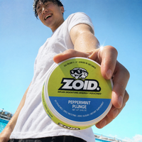 A man in a white t-shirt holds a round container of Zoid Peppermint Plunge energy pouches up close against a bright blue sky and pool background.