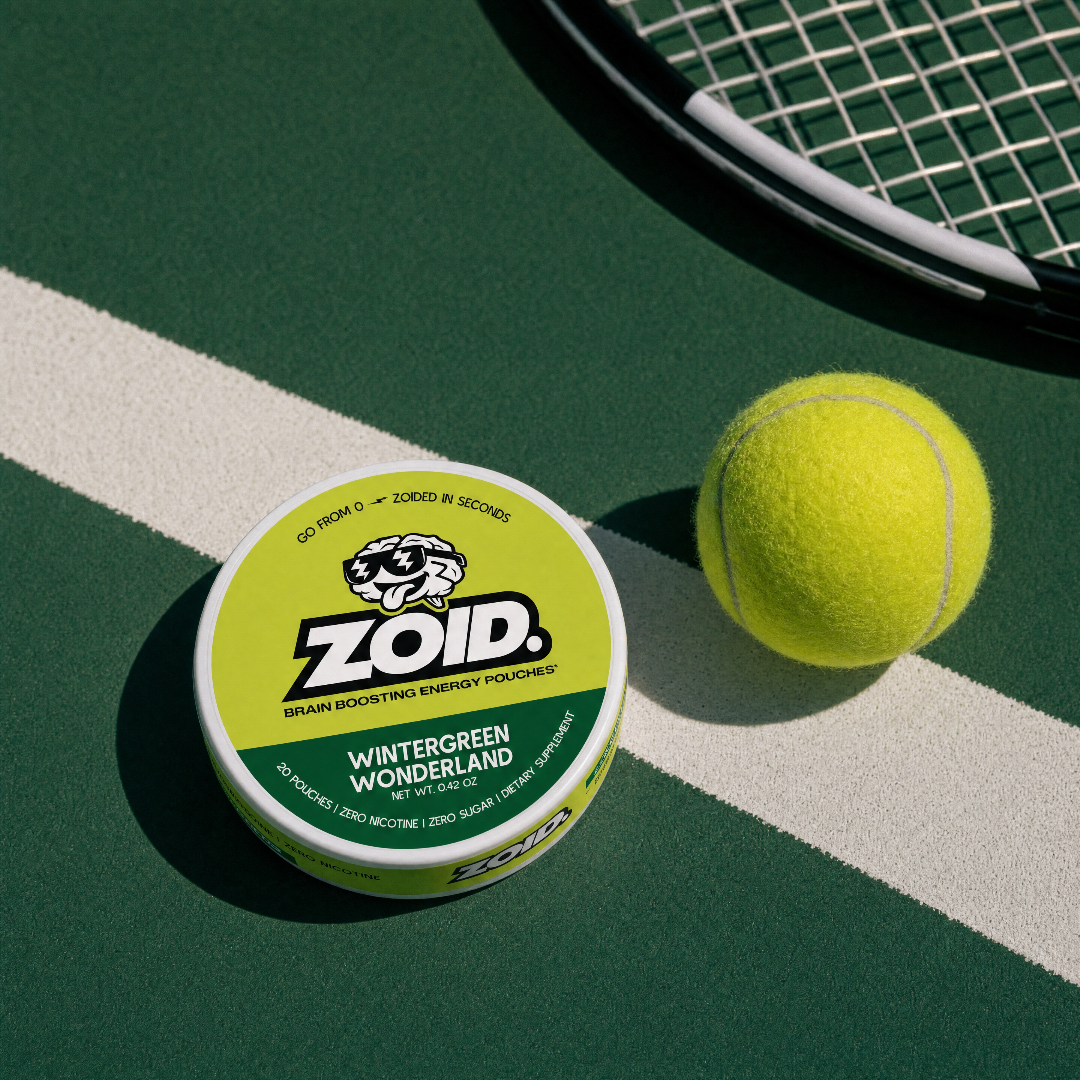 A Zoid Wintergreen Wonderland brain boosting energy pouch can sits on a green tennis court next to a yellow tennis ball, with part of a tennis racket visible in the upper right.